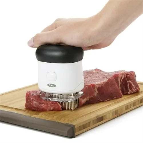 Attendrisseur à viande Oxo en acier inoxydable avec poignée antidérapante utilisé sur un morceau de viande rouge sur planche en bois.